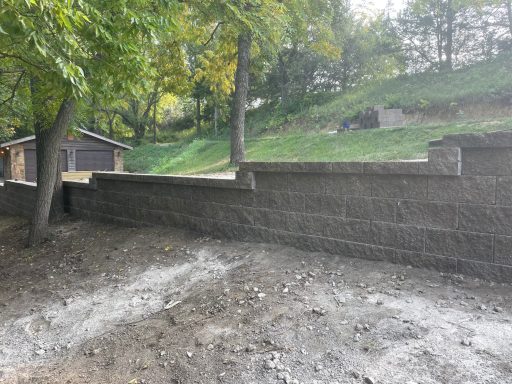 Gray stone wall beside a dirt path, with trees and a nearby building in the background.