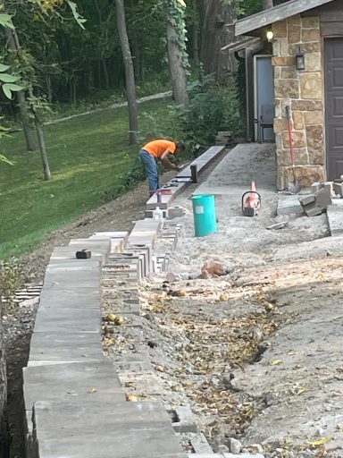 Construction worker in orange shirt working on a stone wall near a house and garden.