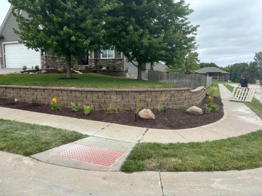 Landscape featuring a stone wall, flower beds, and a curving concrete pathway.