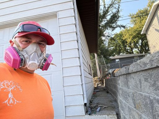 Person wearing a pink mask and sunglasses, standing beside a house under bright sunlight.